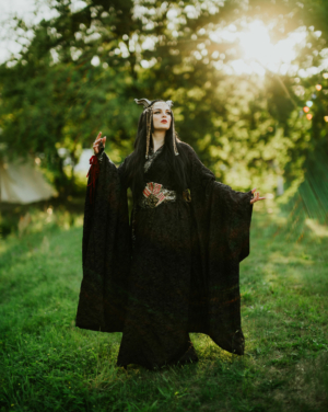 Eine mystische Darstellerin in einem schwarzen Gewand mit aufwendigen Accessoires steht auf einer Wiese beim Elbenwald Festival. Die Sonne scheint durch die Bäume und schafft eine magische Atmosphäre, die das kreative und fantasievolle Ambiente des Festivals unterstreicht.
