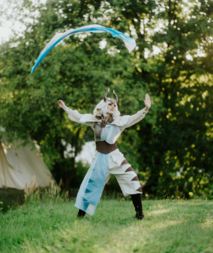 Eine kostümierte Person mit Hörnern und einem fantasievollen Outfit führt eine dynamische Tanz- und Jonglierperformance auf der Wiese des Elbenwald Festivals durch, umgeben von grünen Bäumen und Zelten im Hintergrund.