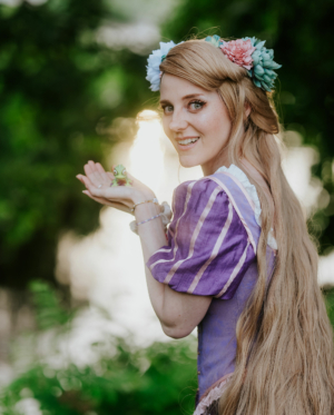 Eine lächelnde Frau mit langen, blonden Haaren und einem Blumenkranz hält einen kleinen Frosch in ihren Händen. Sie trägt ein lila Kostüm mit Streifen und steht vor einer grünen, natürlichen Kulisse, die eine fröhliche Festivalatmosphäre vermittelt.