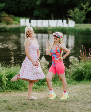 Zwei Frauen in bunten Outfits stehen am Ufer eines Gewässers beim Elbenwald Festival. Im Hintergrund ist das Festival-Logo sichtbar. Die Szenerie strahlt eine fröhliche, festliche Atmosphäre aus, passend zur nerdigen Mischung aus Musik und Convention.