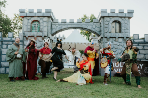 Bunte Gruppe von Cosplayern in fantasievollen Kostümen vor einer mittelalterlichen Kulisse beim Elbenwald Festival, das Musik und Nerdkultur vereint. Die Charaktere repräsentieren verschiedene Genres und Geschichten, die die Besucher begeistern.