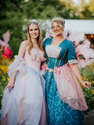 Zwei Frauen in fantasievollen Kostümen posieren fröhlich auf dem Elbenwald Festival vor einer blühenden Kulisse. Die eine trägt ein rosa, glitzerndes Kleid, während die andere in einem blauen Kleid mit Federdetails strahlt. Ein perfekter Moment der Kreativität und Freude.