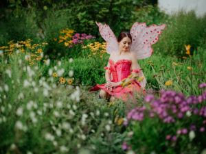 Eine Frau in einem pinken Kostüm mit großen, durchsichtigen Flügeln sitzt in einem bunten Blumenfeld beim Elbenwald Festival. Die lebhaften Farben der Blumen und das fantasievolle Outfit schaffen eine magische, festliche Atmosphäre.