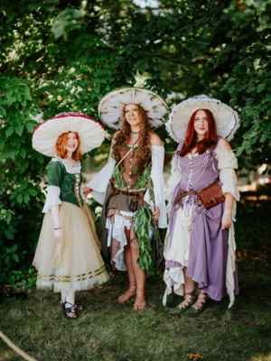 Drei Frauen in fantasievollen Kostümen mit großen, dekorativen Hüten stehen lächelnd vor einer grünen Kulisse. Die Outfits sind inspiriert von Märchen und Fantasy, passend zum Elbenwald Festival, das Musik und Nerd-Kultur vereint.
