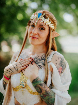 Eine cosplayende Frau mit langen, blonden Haaren und einem goldenen Diadem steht im Freien. Sie trägt ein elegantes, weißes Kostüm und hat kunstvolle Tattoos auf ihren Armen. Der Hintergrund ist unscharf und zeigt grüne Bäume, die eine festliche Atmosphäre schaffen.