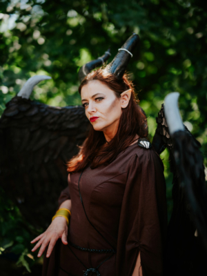 Frau mit Hörnern und schwarzen Flügeln in einem braunen Kostüm, posiert im Freien während des Elbenwald Festivals. Die Szene fängt die kreative Atmosphäre des Festivals ein, das Musik und Fantasy-Elemente vereint.