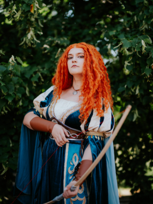 Eine cosplaying Frau mit roten Locken trägt ein elegantes, blaues Kleid und hält einen Bogen in der Hand, umgeben von grünen Blättern. Die Szene spiegelt die kreative Atmosphäre des Elbenwald Festivals wider, das Musik und Nerd-Kultur vereint.