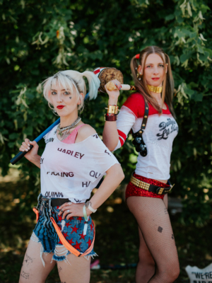 Zwei Frauen in kreativen Harley Quinn Kostümen posieren selbstbewusst vor einem grünen Hintergrund beim Elbenwald Festival. Die Outfits sind farbenfroh und detailreich, mit Accessoires, die den Charakteren aus Comics und Filmen nachempfunden sind.