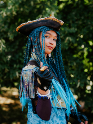 Junge Person mit blauen Zöpfen und Piratenhut, die auf dem Elbenwald Festival posiert. Sie trägt ein kreatives Kostüm mit Glitzerdetails und Accessoires, umgeben von grüner Natur. Ausdruck von Freude und Begeisterung für die Nerdkultur.
