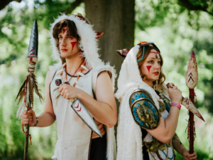 Zwei kostümierte Teilnehmer des Elbenwald Festivals stehen nebeneinander im Freien. Der Mann trägt eine Federkopfbedeckung und hält einen Speer, während die Frau in einem aufwendigen Outfit mit Schmuck und ebenfalls einem Speer posiert.