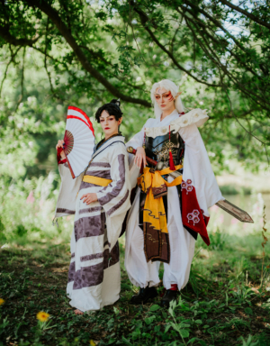 Zwei Cosplayer in fantasievollen Kostümen stehen unter einem Baum auf dem Elbenwald Festival. Die erste trägt ein traditionelles Outfit mit Fächer, während die zweite eine beeindruckende Rüstung und ein Schwert präsentiert. Bunte Natur im Hintergrund.