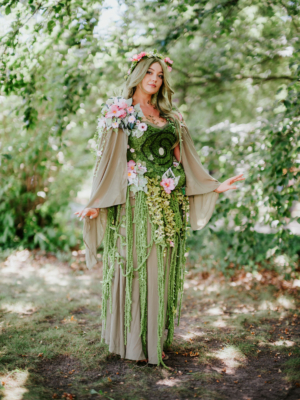 Eine kostümierte Teilnehmerin des Elbenwald Festivals steht in einem grünen, blumengeschmückten Outfit zwischen Bäumen. Ihr Look kombiniert Natur- und Fantasyelemente, perfekt für die kreative Atmosphäre des Festivals.