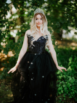 Eine junge Frau in einem glamourösen schwarzen Kleid mit funkelnden Details und einer Krone posiert im Freien, umgeben von grünen Bäumen und einem Hauch von Glitzer, passend zur kreativen Atmosphäre des Elbenwald Festivals.