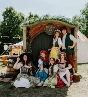 Gruppenszene von Cosplayern in Fantasiekostümen vor einem Hobbit-Eingang beim Elbenwald Festival. Die Charaktere sind inspiriert von "Der Herr der Ringe", umgeben von bunten Dekorationen und einem festlichen Ambiente.