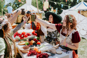 Gruppe von Frauen in fantasievoller Kleidung hebt ihre Tassen zum Anstoßen, umgeben von einem festlich gedeckten Tisch mit frischen Früchten und einer Torte. Im Hintergrund sind bunte Wimpel und Zelte des Elbenwald Festivals sichtbar.