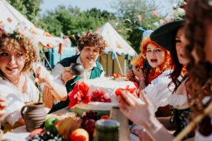 Eine Gruppe von verkleideten Festivalbesuchern genießt ein festliches Mahl beim Elbenwald Festival. Bunte Kostüme, eine festliche Torte und eine fröhliche Atmosphäre schaffen eine lebendige Szene im Freien, umgeben von bunten Fahnen und Zelten.
