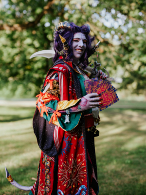 Eine cosplayende Person mit lila Haaren und Hörnern hält bunte Karten in der Hand. Sie trägt ein auffälliges, farbenfrohes Kostüm mit verschiedenen Mustern und Accessoires. Hintergrund mit grünem Gras und Bäumen, passend zur lebhaften Atmosphäre des Elbenwald Festivals.