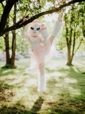 Ein Tänzer in einem niedlichen Katzenkostüm mit rosa Tüllrock und weißen Stiefeln, der auf einem Bein balanciert und an einem Baum hängt. Die Szene ist umgeben von grünem Gras und Sonnenlicht, perfekt für das Elbenwald Festival.