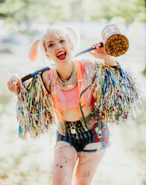 Frau mit buntem Outfit und Glitzerjacke, die fröhlich mit einem großen Hammer posiert. Im Hintergrund ist eine grüne Wiese zu sehen, die festliche Atmosphäre des Elbenwald Festivals unterstreicht. Ein kreatives und nerdiges Event voller Spaß und Musik.