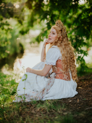 Eine Frau mit langen, lockigen Haaren sitzt im Freien auf dem Boden, trägt ein elegantes, historisches Kleid und eine Blumenkrone. Sie blickt über die Schulter, umgeben von grüner Natur, während die Sonne sanft auf die Szenerie scheint.