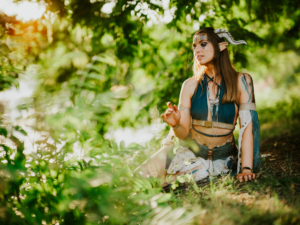 Eine cosplaying Frau mit Elfenohren und fantasievoller Kleidung sitzt im Grünen beim Elbenwald Festival, umgeben von Bäumen und Sonnenlicht, und verkörpert die kreative Atmosphäre des Festivals, das Musik und Nerdkultur vereint.