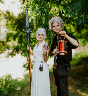 Zwei Cosplayer auf dem Elbenwald Festival, gekleidet in fantasievolle Kostüme. Die weibliche Figur trägt ein weißes Kleid und hält einen magischen Gegenstand, während der männliche Cosplayer eine Laterne und einen Speer präsentiert. Grüne Natur im Hintergrund.