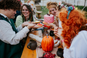 Eine Gruppe von Menschen in fantasievoller Kleidung sitzt an einem Tisch, umgeben von Kürbissen und bunten Süßigkeiten. Sie halten eine Torte und Gebäck in den Händen und lachen, während sie die festliche Atmosphäre des Elbenwald Festivals genießen.