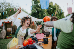 Lächelnde Teilnehmer des Elbenwald Festivals in mittelalterlicher Kleidung feiern mit bunten Speisen und Getränken an einem festlich dekorierten Tisch, umgeben von Zelten und farbigen Fähnchen. Die Atmosphäre ist fröhlich und einladend, ideal für Nerds und Fantasy-Fans.