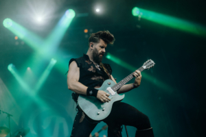 dArtagnan Ein Musiker mit einem hellblauen E-Gitarre spielt leidenschaftlich auf der Bühne beim Elbenwald Festival, umgeben von grünem Licht und einer energiegeladenen Atmosphäre. Die Szene fängt die Dynamik und den Spaß des Festivals perfekt ein.