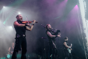 dArtagnan Auf der Bühne des Elbenwald Festivals spielen Musiker mit Geige und Dudelsack. Die Atmosphäre ist lebhaft und mystisch, umgeben von buntem Licht und Nebel, während die Band das Publikum begeistert.