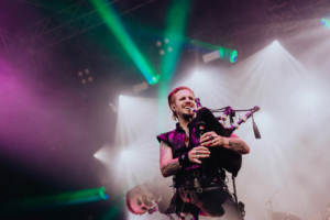 dArtagnan Ein Musiker spielt mit Leidenschaft Dudelsack auf der Bühne des Elbenwald Festivals, umgeben von bunten Lichtstrahlen und Nebel, während die Menge begeistert zuschaut. Die Atmosphäre vereint Musik und Nerdkultur in einem einzigartigen Erlebnis.