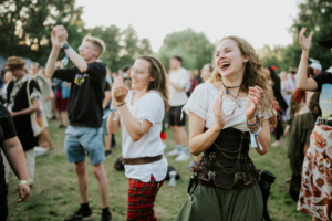Festivalbesucher feiern begeistert beim Elbenwald Festival, gekleidet in fantasievolle Kostüme. Die fröhliche Stimmung und das gemeinsame Klatschen schaffen eine lebendige Atmosphäre inmitten der Natur. Ein unvergessliches Erlebnis für Nerds und Musikliebhaber.