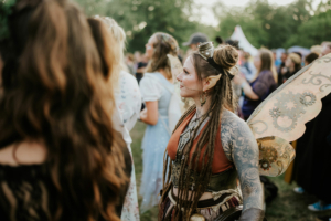 Eine Frau mit Elfenohren und aufwendigen Tattoos trägt ein fantasievolles Kostüm mit mechanischen Flügeln beim Elbenwald Festival, umgeben von anderen Festivalbesuchern in einem lebhaften Freiluftbereich.