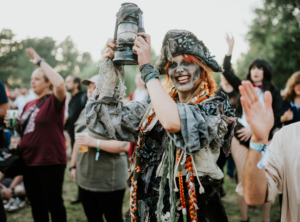 Eine fröhliche Festivalbesucherin in fantasievoller Piratenkostümierung hält eine Laterne hoch, umgeben von einer feiernden Menge beim Elbenwald Festival. Die Atmosphäre ist lebhaft und zeigt die kreative Vielfalt der Teilnehmer.
