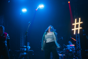 #zweiraumsilke Auf der Bühne des Elbenwald Festivals performt eine Künstlerin leidenschaftlich, während das Bühnenlicht in Blau erstrahlt. Im Hintergrund sind Musiker am Schlagzeug und Keyboard zu sehen, die die energiegeladene Atmosphäre unterstützen.