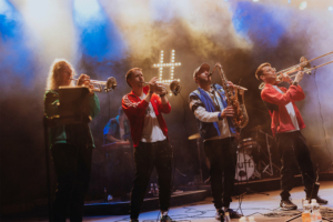 #zweiraumsilke Vier Musiker auf der Bühne des Elbenwald Festivals spielen Blasinstrumente inmitten von Nebel und bunten Lichtern. Die energiegeladene Performance zieht die Aufmerksamkeit des Publikums auf sich und schafft eine lebhafte Festivalatmosphäre.