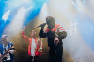 #zweiraumsilke Auf der Bühne des Elbenwald Festivals performt eine Band mit einem Sänger, der ein Mikrofon hält, während ein Trompeter und ein Saxophonist im Hintergrund aktiv sind. Bunte Lichter und Nebel schaffen eine lebendige Festivalatmosphäre.