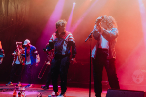 #zweiraumsilke Auf der Bühne des Elbenwald Festivals performt eine Band mit mehreren Musikern in sportlicher Kleidung. Die Lichter blitzen in verschiedenen Farben, während die Künstler mit Mikrofonen energisch auftreten und die Menge begeistern.
