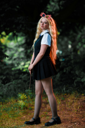 Eine junge Frau mit langen, blonden Haaren trägt ein stylisches Outfit mit einem schwarzen Rock und einer weißen Bluse. Sie hat eine besondere Brille auf und hält einen Zauberstab, während sie in einem grünen, bewaldeten Bereich des Elbenwald Festivals posiert.