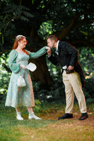 Ein Paar in fantasievoller Kleidung posiert auf dem Elbenwald Festival, während der Mann der Frau die Hand küsst. Die Frau trägt ein elegantes, mintgrünes Kleid und der Mann einen schwarzen, kunstvoll verzierten Frack. Hintergrund aus Bäumen und Gras.