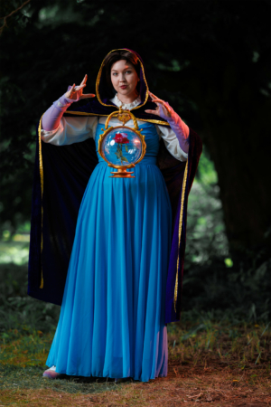 Eine Cosplayerin in einem blauen Kleid und einem schwarzen Umhang hält eine magische Glaskugel mit einer Rose. Der Hintergrund zeigt eine grüne, bewaldete Umgebung, die zur Atmosphäre des Elbenwald Festivals beiträgt.