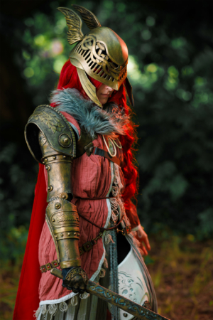 Kostümierte Person mit rotem Haar und goldener Rüstung auf dem Elbenwald Festival, umgeben von grüner Natur. Die Figur hält ein Schwert und trägt einen gefiederten Helm, was die Verbindung zu Fantasy und Rollenspielen unterstreicht.