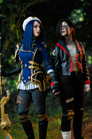 Cosplay-Duo auf dem Elbenwald Festival, verkleidet als Charaktere aus einem beliebten Videospiel. Die Frau trägt eine blaue Uniform mit goldenen Akzenten, während der Mann in einer schwarzen Jacke mit roten Details und Bandagen auftritt. Grüner Hintergrund.